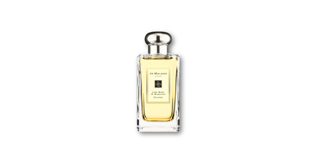 Jo Malone Lime Basil & Mandarin cologne bottle surrounded by fresh limes, basil leaves, and mandarin oranges on a wooden table, suggesting a zesty, herbaceous unisex fragrance.