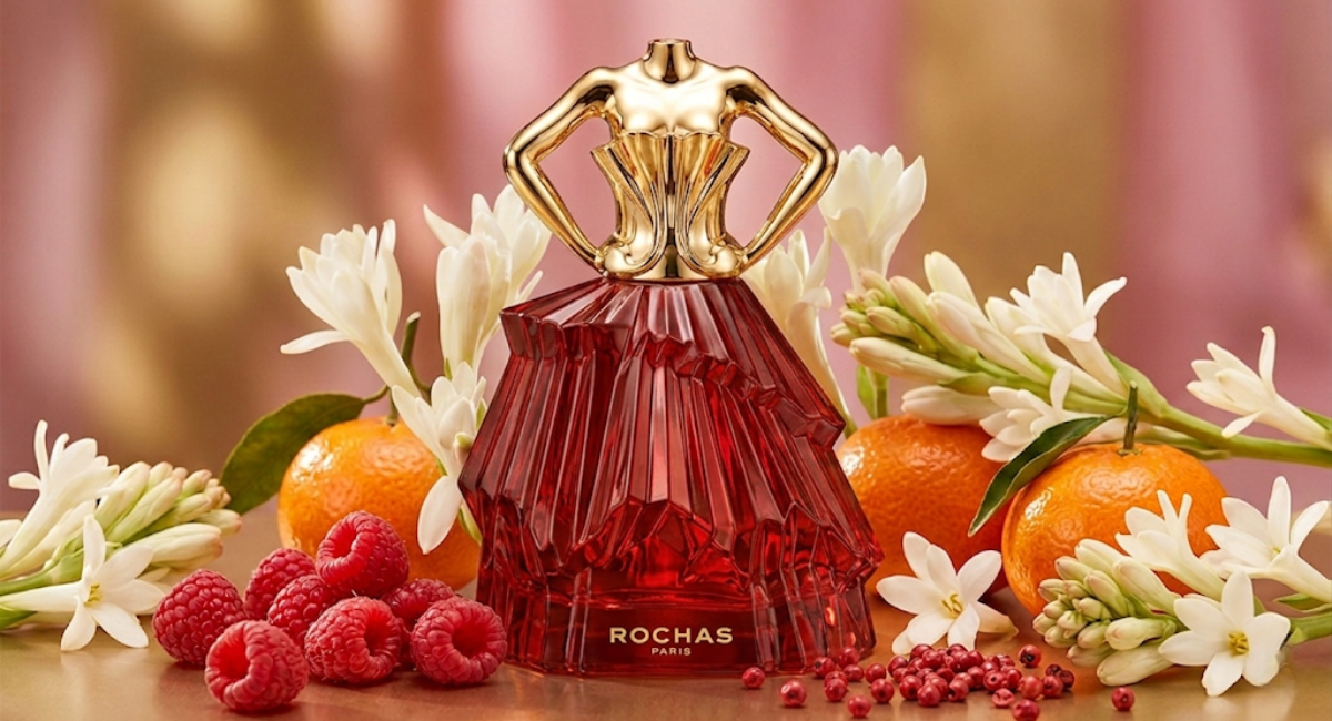 Rochas Audace Le Parfum 2026 bottle – golden trophy design with faceted glass body, surrounded by raspberries, mandarins, and tuberose flowers