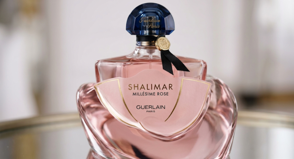 Close-up of Shalimar Millesime Rose bottle showing pink batwing label with gold accents, deep blue faceted cap, and black ribbon with gold medallion seal